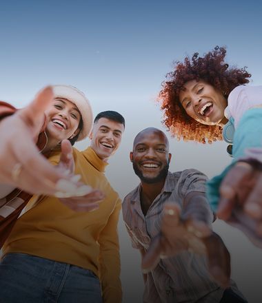 A group of four is smiling and reaching toward the camera from a low-angle view against a clear sky