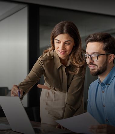 A woman leaning over to assist a man at a desk, both looking at something in front of them and smiling